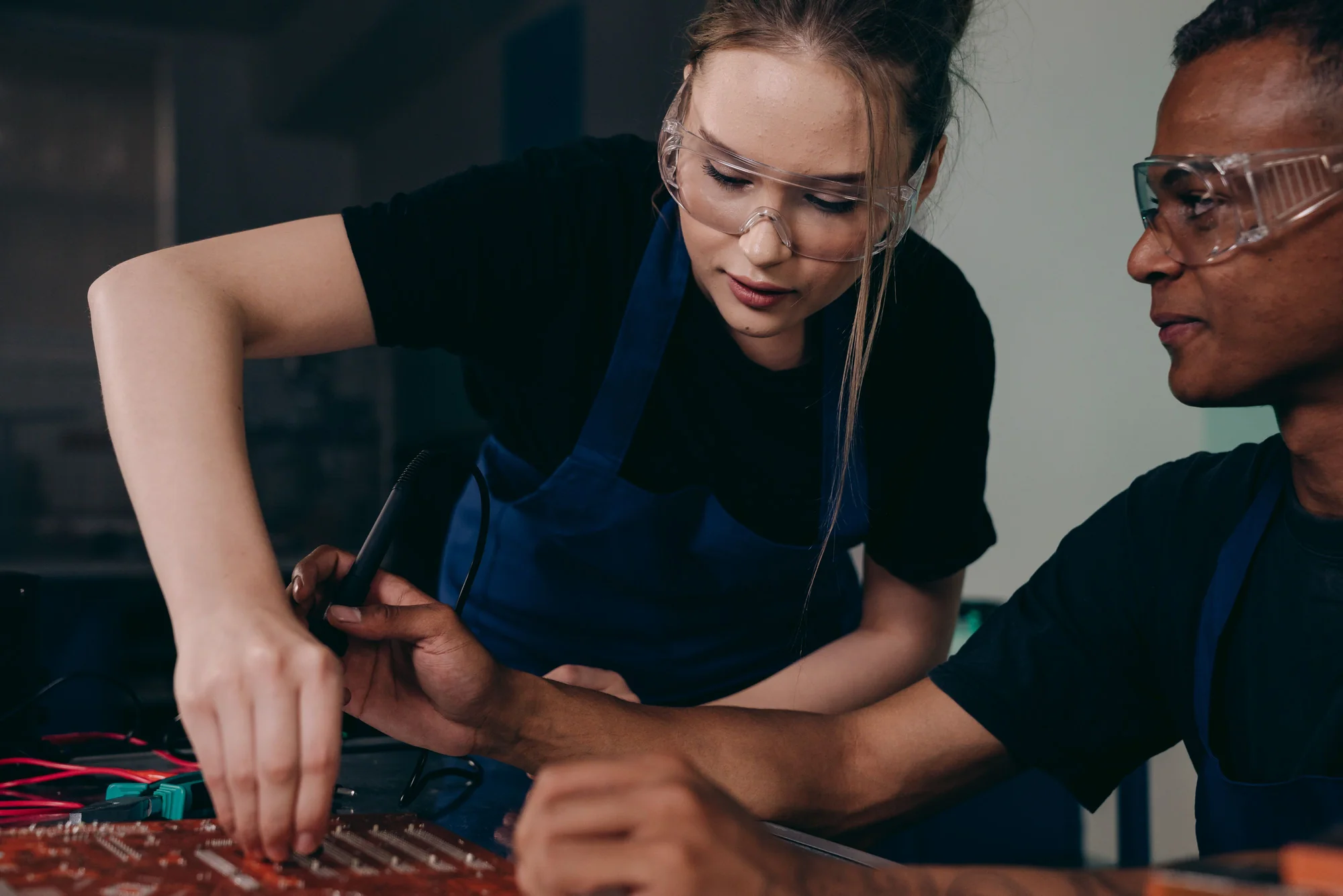 Two people wearing protective eyewear examine an electronic assembly at an engineering workbench
