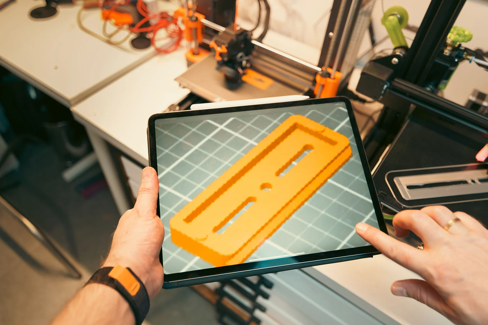 A tablet displays a three-dimensional prototype model beside workshop 3D printers